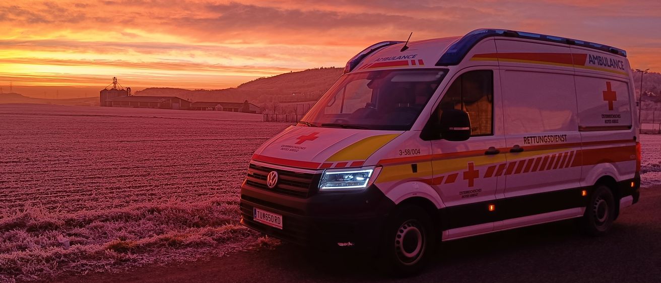 Ein Krankenwagen mit der Nummer 3-58-004 ist auf einer Straße geparkt unter einem Sonnenuntergang mit Blick auf eine Fabrik und Hügel in der Ferne.