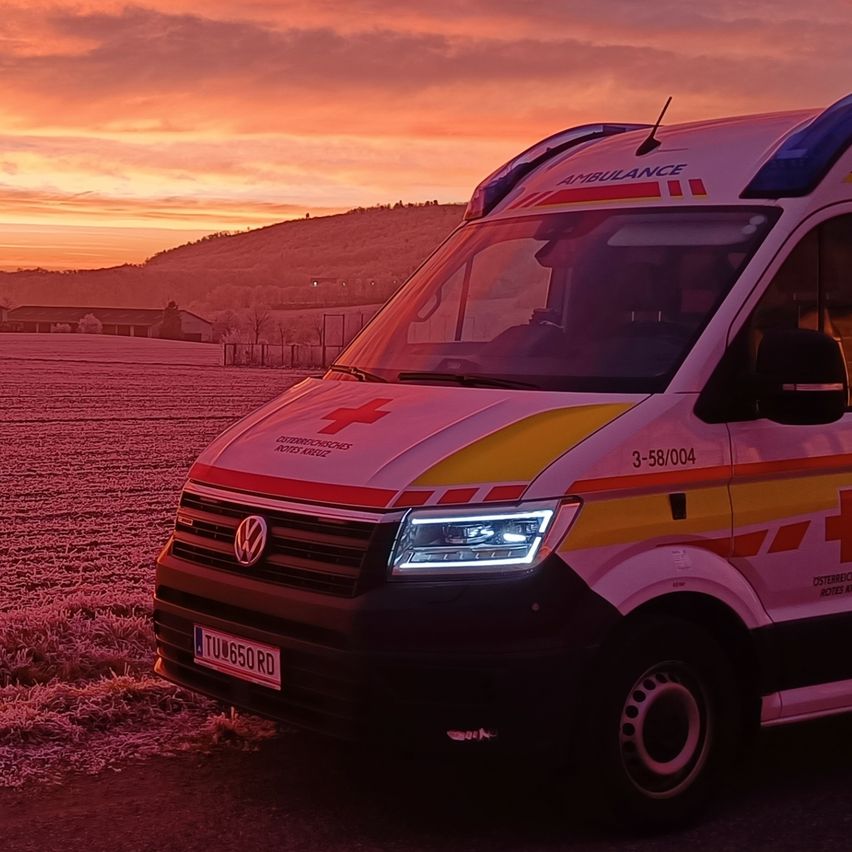 Ein Krankenwagen mit der Nummer 3-58-004 ist auf einer Straße geparkt unter einem Sonnenuntergang mit Blick auf eine Fabrik und Hügel in der Ferne.