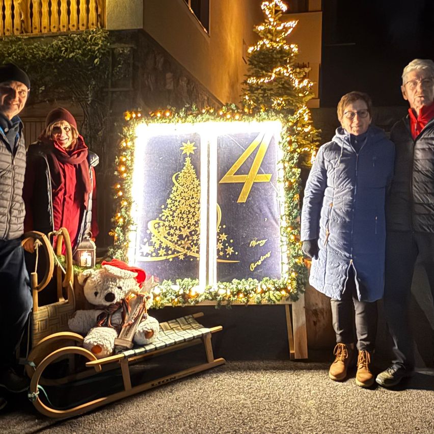 Vier Personen stehen vor einem festlich geschmückten Weihnachtsbaum, der mit Lichtern und Ornamenten verziert ist. Einer sitzt in einem Schlitten mit einem Teddybären, während die anderen für die Kamera lächeln.