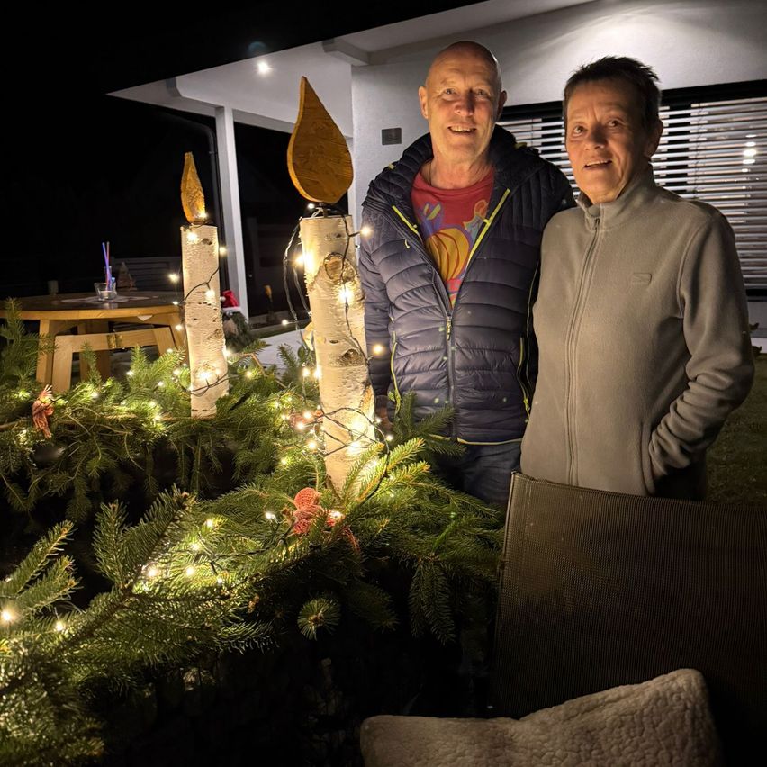 Two individuals, a man and a woman, stand in front of a decorated outdoor area with lights and Christmas trees. They are smiling and posing for a picture.