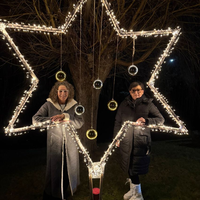 Zwei Frauen stehen unter einer sternförmigen Lichtinstallation mit hängenden Ornamenten, eine mit Brille und die andere mit einem Lächeln, vor einem Baum.