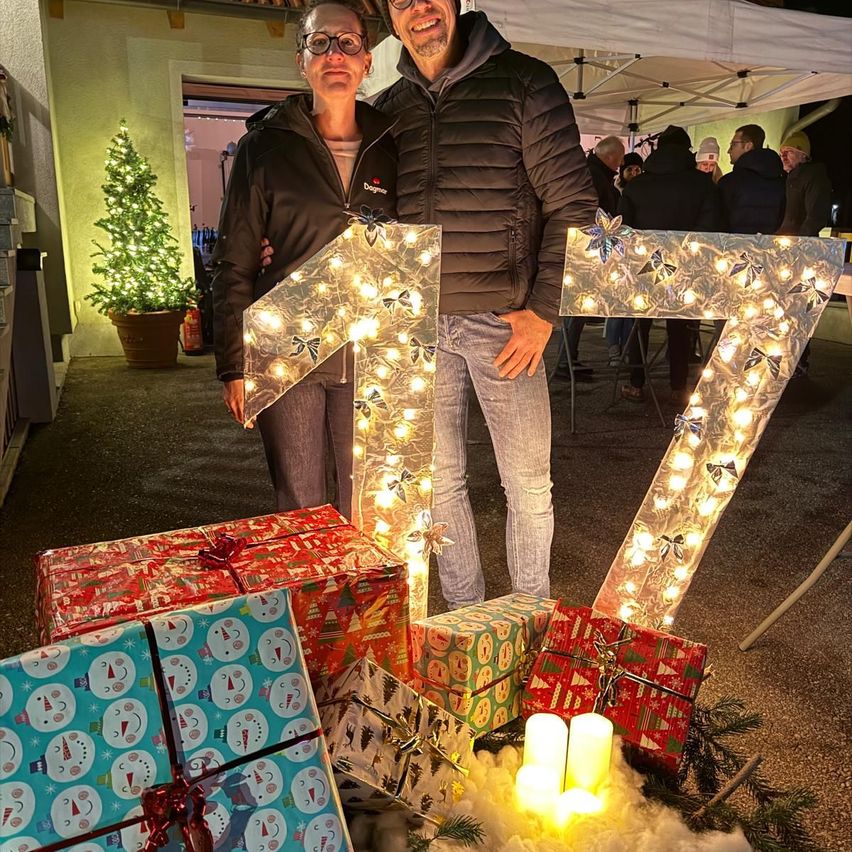 Ein Mann und eine Frau stehen neben einer aus Lichtern bestehenden Nummer 17, umgeben von Weihnachtsgeschenken. Sie lächeln und posieren für ein Foto. Hinter ihnen versammeln sich Leute unter einem Zelt.