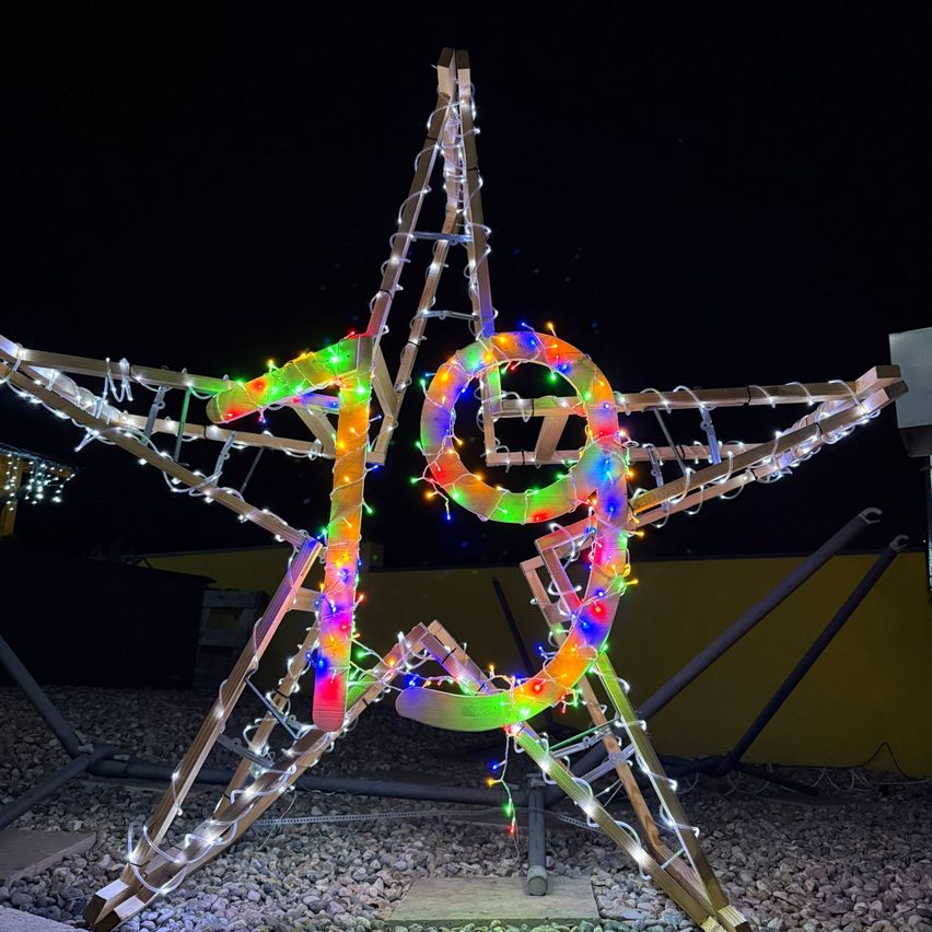 Eine sternförmige Struktur aus Holz ist mit bunten Lichtern beleuchtet. Der Stern steht auf einer felsigen Oberfläche, mit einer gelben Wand im Hintergrund.