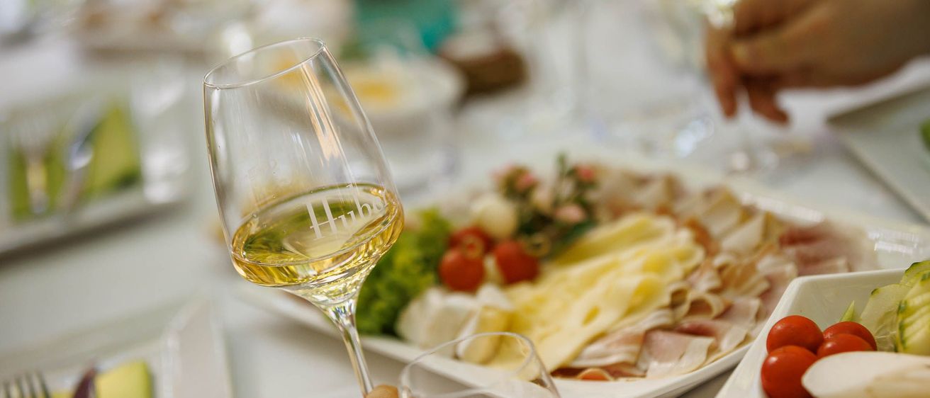 Eine Hand hält ein Weinglas mit der Aufschrift 'Hub' mit Weißwein, vor einem Tisch mit einem Käsetablett und einem unscharfen Hintergrund.