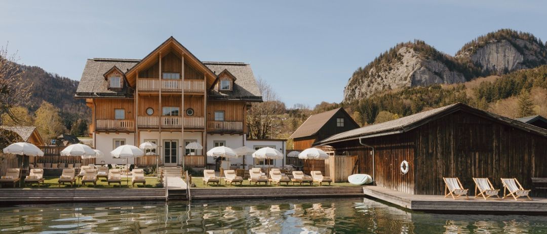 Ein Holzresort mit mehreren Gebäuden, darunter ein Balkon, und Liegestühlen unter Sonnenschirmen. Das Resort liegt in der Nähe eines Sees mit Reflexionen der Gebäude.