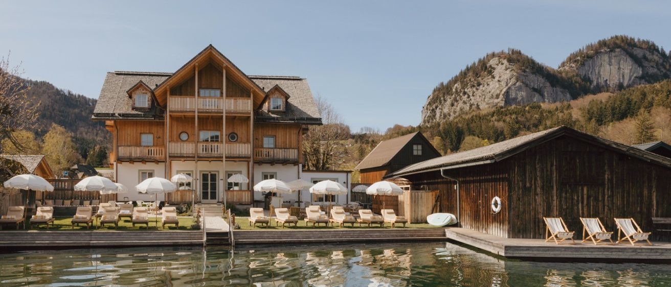 Ein Holzresort mit mehreren Gebäuden, darunter ein Balkon, und Liegestühlen unter Sonnenschirmen. Das Resort liegt in der Nähe eines Sees mit Reflexionen der Gebäude.