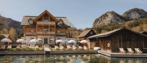 Ein Holzresort mit mehreren Gebäuden, darunter ein Balkon, und Liegestühlen unter Sonnenschirmen. Das Resort liegt in der Nähe eines Sees mit Reflexionen der Gebäude.