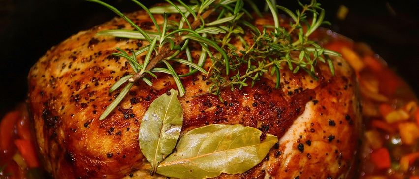 A close-up of a roasted chicken dish, garnished with rosemary and bay leaves, showing its browned and crispy skin with herbs and spices sprinkled on top.