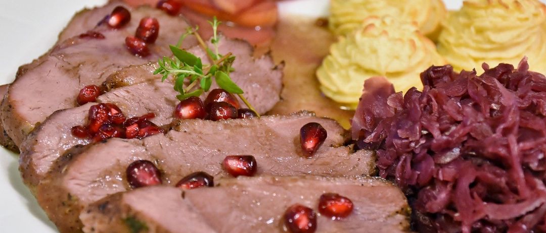 Ein weißer Teller enthält Scheiben von gebratenem Fleisch mit roten Granatapfelsamen, gelber Kartoffelpüree und rotem Kraut. Es gibt einen Kräuterzweig auf dem Fleisch.