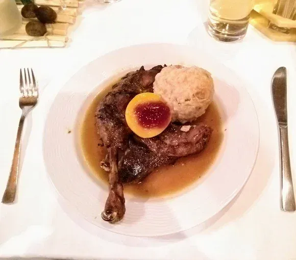 Ein weißer Teller mit einem Lammkeule mit Soße, einer Kartoffelknödel und einem halben Pfirsich. Ein Messer und eine Gabel sind auf dem Tisch.