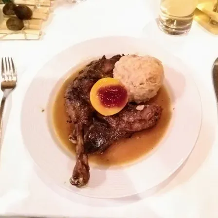 Ein weißer Teller mit einem Lammkeule mit Soße, einer Kartoffelknödel und einem halben Pfirsich. Ein Messer und eine Gabel sind auf dem Tisch.