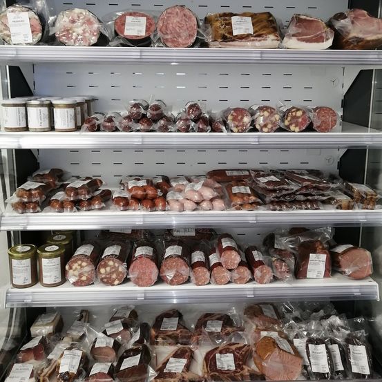 Eine gekühlte Vitrine mit mehreren Regalen verschiedener Fleischsorten, darunter Würste, Salami und andere Wurstwaren, alles in Plastik verpackt und beschriftet.