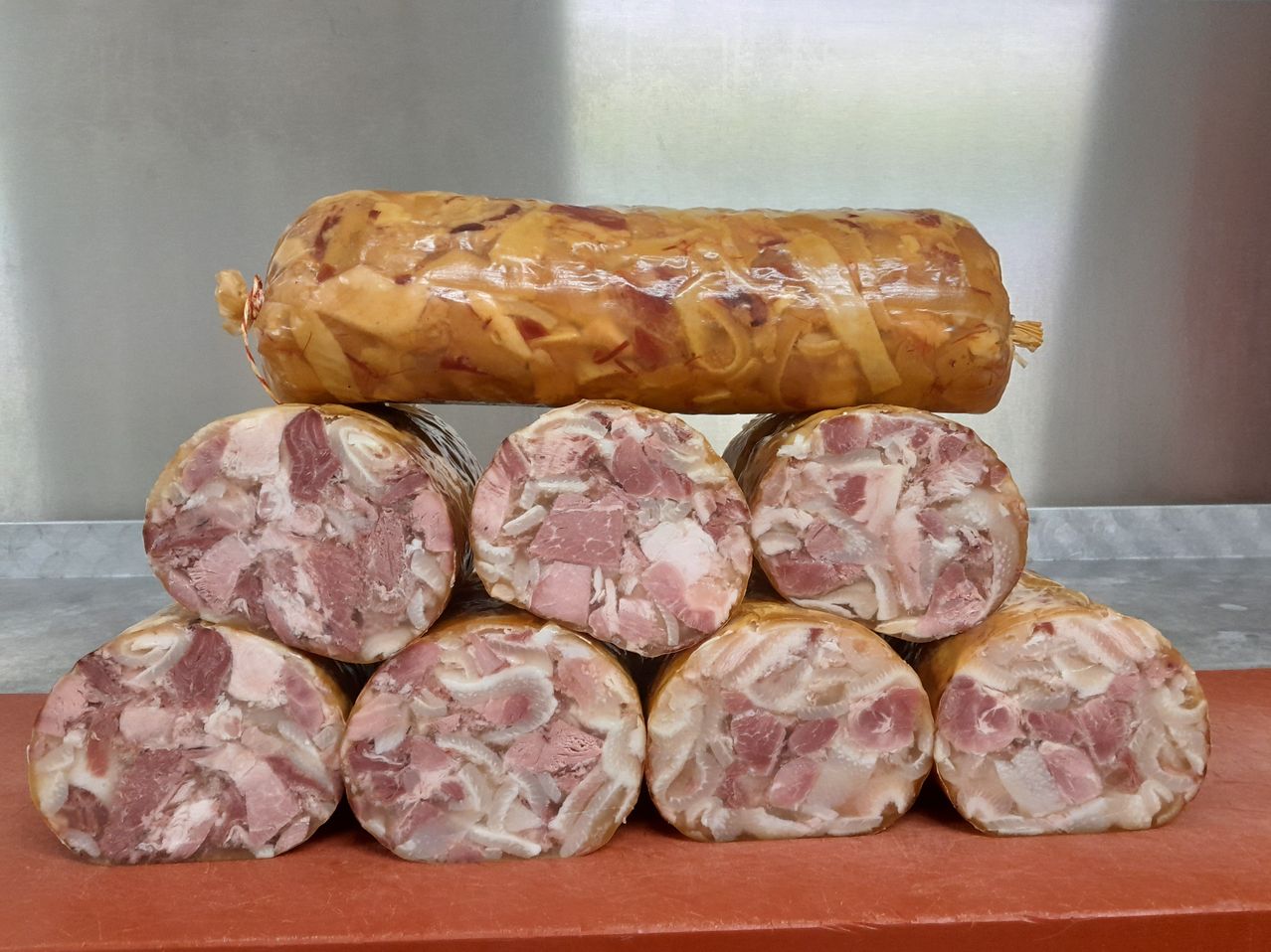 Mehrere Würste, einige geschnitten und andere ganz, sind auf einer braunen Oberfläche ausgestellt. Die geschnittenen Würste haben eine Mischung aus rosa und weißem Fleisch. Ein größerer Wurst ist in Plastik eingewickelt und obenauf gelegt.