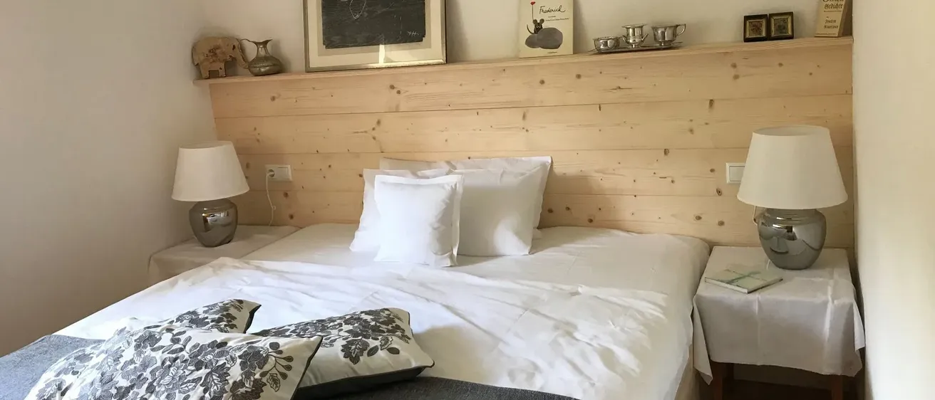 Ein Schlafzimmer mit einem Holzkopfteil hat ein Bett mit weißen Laken und Kissen. Zwei Lampen und ein Buch stehen auf den Nachttischen. Ein Bilderrahmen und Dekorationsartikel schmücken die Wand über dem Bett.
