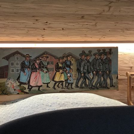 Ein Wandbild im Schlafzimmer zeigt eine Gruppe von Menschen in traditioneller Kleidung, die in einer Reihe gehen. Das Wandbild zeigt Gebäude, einen Baum und ein helles Licht.