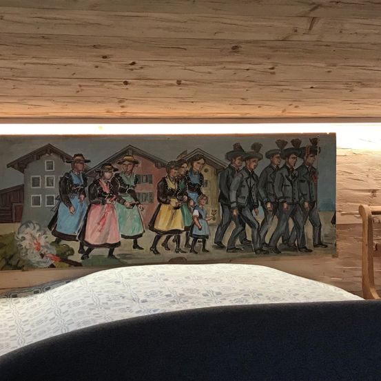 Ein Wandbild im Schlafzimmer zeigt eine Gruppe von Menschen in traditioneller Kleidung, die in einer Reihe gehen. Das Wandbild zeigt Gebäude, einen Baum und ein helles Licht.
