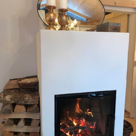 Ein Kamin mit brennendem Feuer im Inneren, Kerzen darauf und einem Spiegel, der den Raum reflektiert. Holzscheite sind links gestapelt.