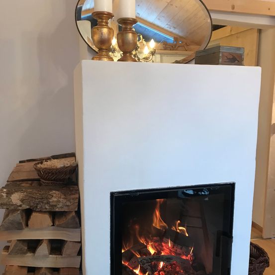 Ein Kamin mit brennendem Feuer im Inneren, Kerzen darauf und einem Spiegel, der den Raum reflektiert. Holzscheite sind links gestapelt.