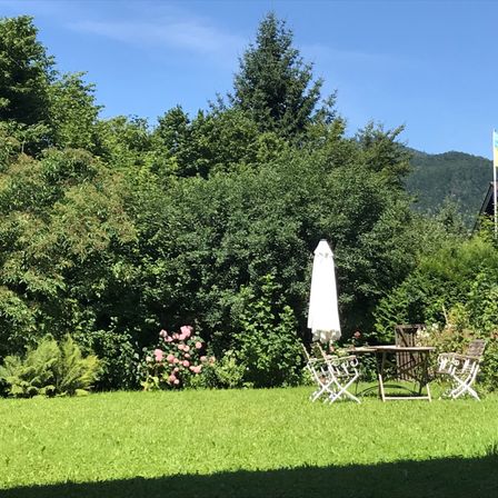 Ein Garten mit einem Tisch und Stühlen unter einem Schirm, umgeben von Grün und Blumen, mit einer Flagge in der Ferne.