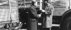 Zwei Männer in Mänteln schütteln sich die Hände vor einem Lastwagen mit der Nummer 781. Ein zweiter LKW im Hintergrund trägt die Aufschrift Alois Schott und GRAZ.