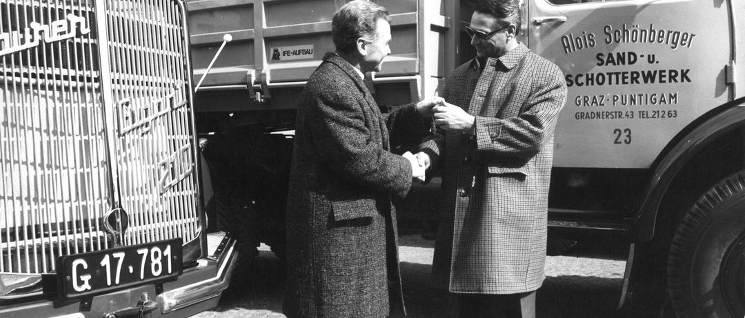 Zwei Männer in Mänteln schütteln sich die Hände vor einem Lastwagen mit der Nummer 781. Ein zweiter LKW im Hintergrund trägt die Aufschrift Alois Schott und GRAZ.