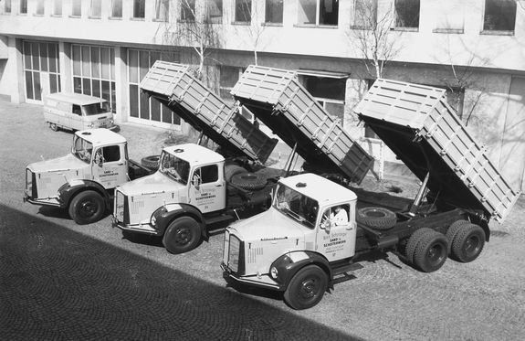 Ein Schwarz-Weiß-Foto zeigt vier Lastwagen mit angehobenen Kippaufbauten. Sie stehen vor einem Gebäude mit vielen Fenstern. Ein Lieferwagen ist ebenfalls geparkt.