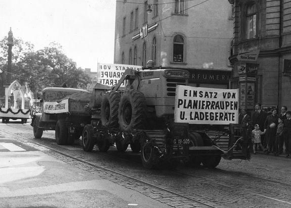 Ein altes Foto eines Traktors in einer Parade, mit Schildern, die 'Einsatz von Planierraupen U. Ladegerate' werben. Leute beobachten.