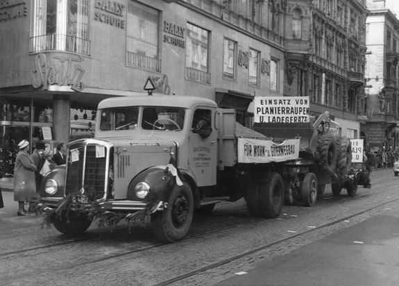Ein alter LKW fährt mit einem Banner und einem Elefanten die Straße entlang. Menschen stehen auf dem Bürgersteig, und ein Gebäude hat ein Schild.
