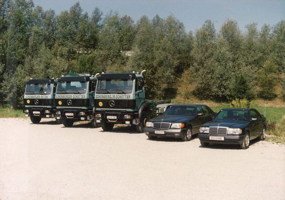 Drei Lastwagen und zwei Autos sind auf einer Straße geparkt, mit grünen Bäumen im Hintergrund. Die Lastwagen haben weiße Schrift auf ihren Seiten.