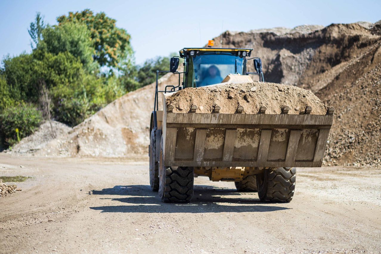 A yellow bulldozer is driving down a dirt road, loaded with soil, with a person inside.