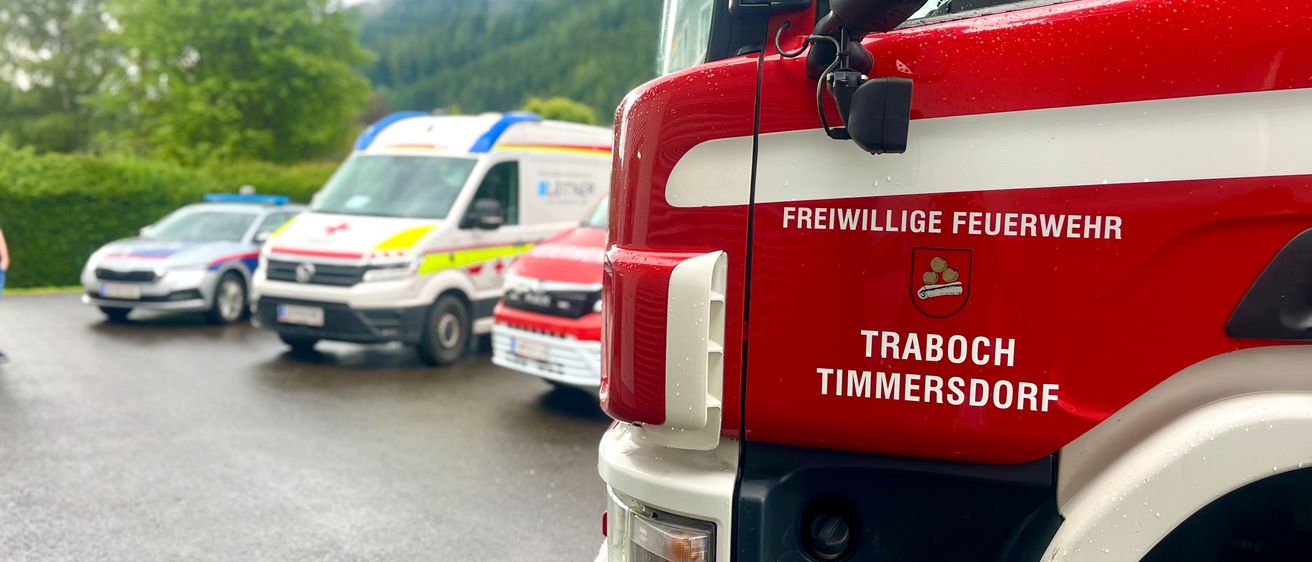 Ein Feuerwehrwagen aus Traboch Timmersdor steht auf einem Parkplatz mit Polizei- und Krankenwagen. Eine Person steht in der Nähe.