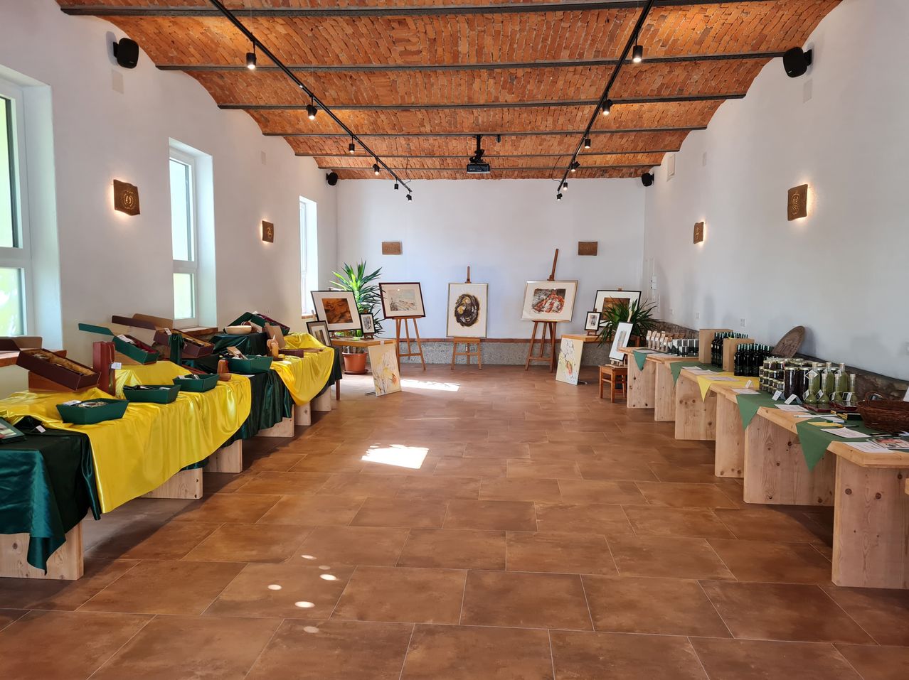 Ein leerer Ausstellungssaal mit braunen Fliesenböden und Ziegeldecke. Kunstwerke sind auf Staffeleien ausgestellt, einige mit gelben Abdeckungen. Holztische mit grünen Abdeckungen sind aufgestellt, und einige Topfpflanzen stehen in den Ecken. Scheinwerfer sind an der Decke montiert.