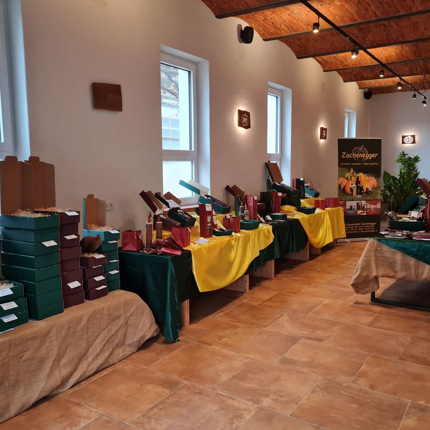 Ein Saal mit gefliesten Böden und mehreren Tischen, die mit gelben Tüchern bedeckt sind. Stapel von Geschenkboxen in Grün und Braun befinden sich auf der linken Seite. Ein Schild für Zachegger ist an der Wand angebracht.