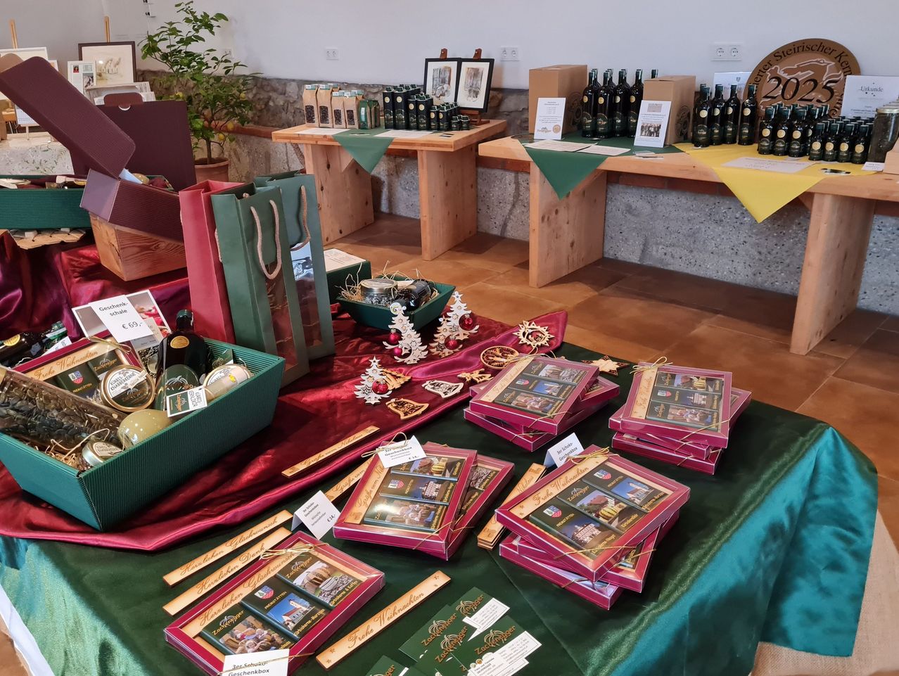 Ein Tisch zeigt verschiedene Geschenkboxen, einige mit Pralinen, andere mit dekorativen Gegenständen. Hinter dem Tisch stehen Holzbänke mit Weinflaschen und Kisten darauf. Die Umgebung umfasst Topfpflanzen und eine Wand mit einem kreisförmigen Schild.