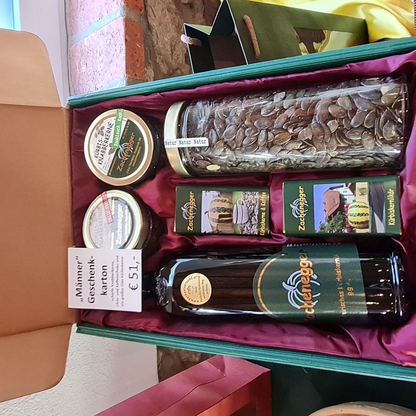 Eine grüne Geschenkbox mit rotem Futter enthält eine große Glaskanne mit Samen, eine Ölflasche und zwei Gläser Marmelade, alles auf einem roten Stoffbett.
