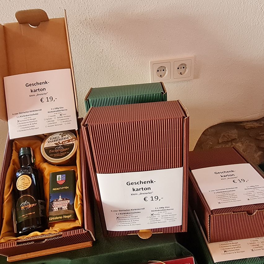 Geöffnete Geschenkboxen mit Etiketten, die 'Geschenk-Karton klein, 'Dreierle', €19' lesen. Jede Box enthält eine Flasche Schnaps, eine Dose und ein Büchlein. Die Flaschen haben Etiketten mit Text und Logos. Hinter den Boxen ist eine Steckdose an einer weißen Wand angebracht.