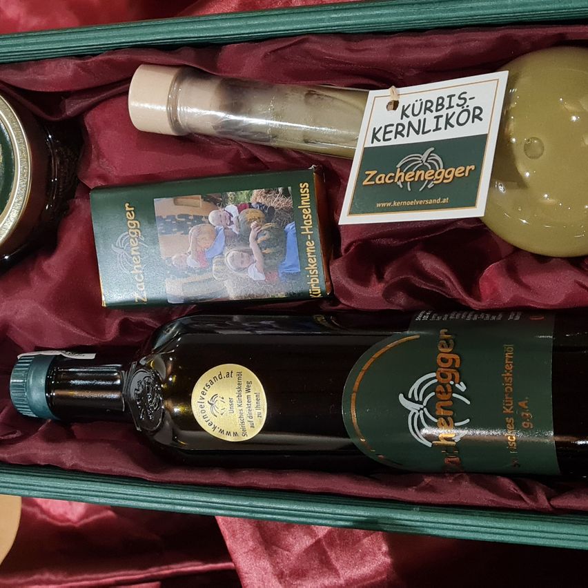 Ein Geschenkset mit einer Flasche Olivenöl, einem kleinen Buch, einer Kernlikör und einem kleinen Glas, alles ordentlich in einer grünen Box auf einem roten Samttuch angeordnet.