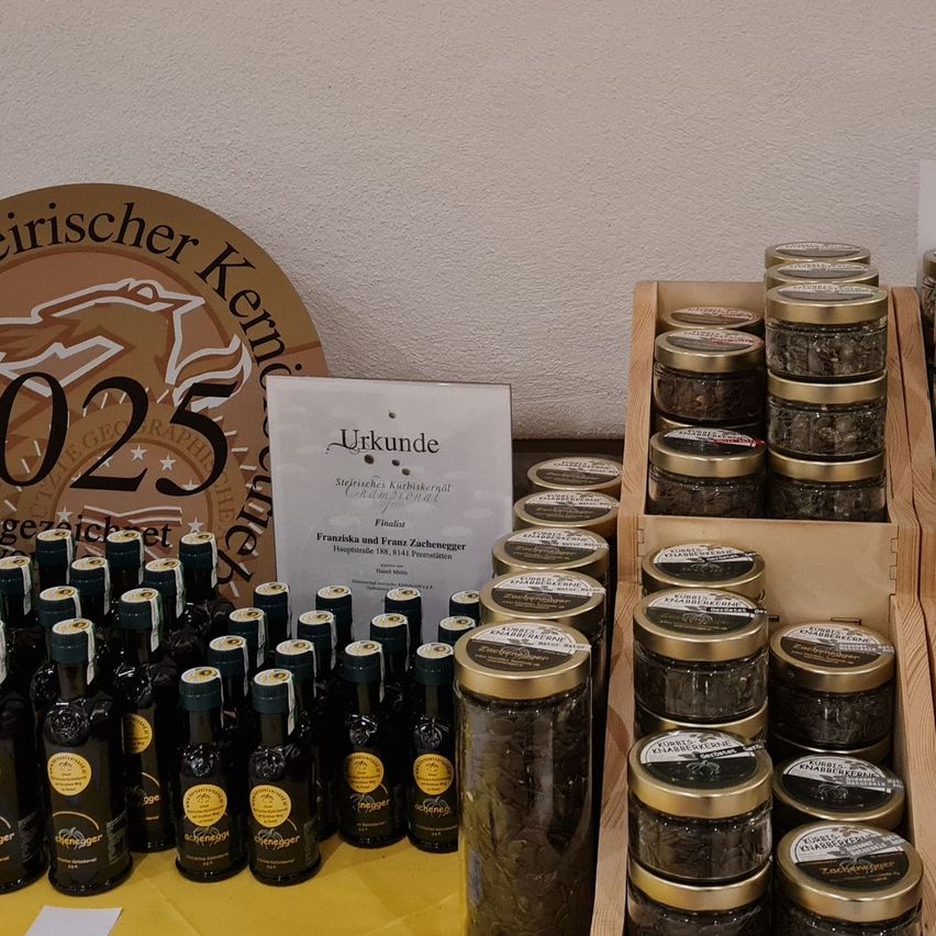 Ein Tisch zeigt mehrere Gläser mit Lebensmittelprodukten, einige mit goldenen Deckeln. Dahinter befindet sich ein Zertifikat mit der Aufschrift 'Urkunde'. Über den Gläsern ist ein rundes Schild mit der Zahl '25' und dem Wort 'Geographie' zu sehen.