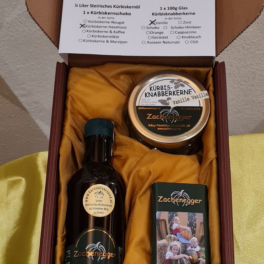 Eine Schachtel enthält eine Flasche Kürbiskernöl, ein Glas Kürbiskern-Nougat und eine kleine Schachtel. Das Etikett erwähnt verschiedene Geschmacksrichtungen und einen direkten Weg zur Kürbis.