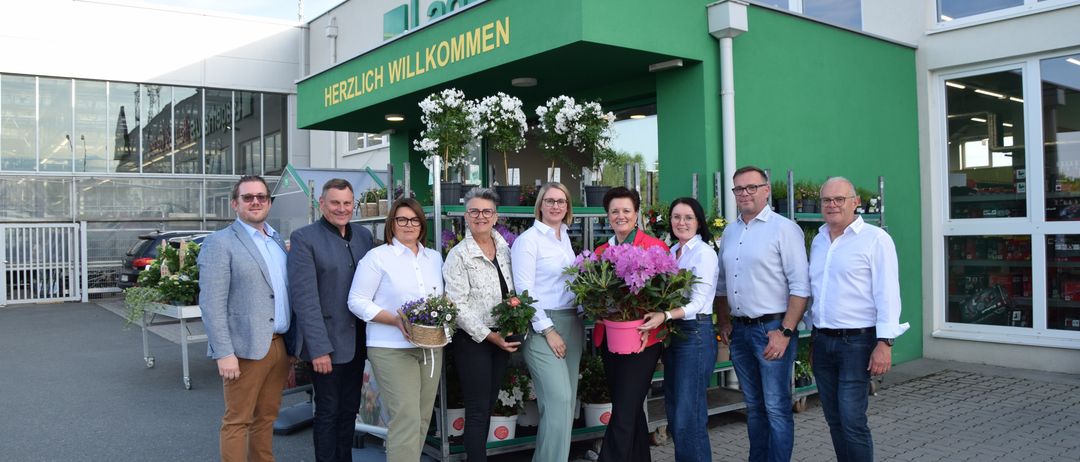 Eine Gruppe von Menschen steht vor einem Blumenladen und lächelt für ein Foto. Sie halten Blumentöpfe und Körbe. Der Laden hat ein grünes Vordach mit weißen Texten.