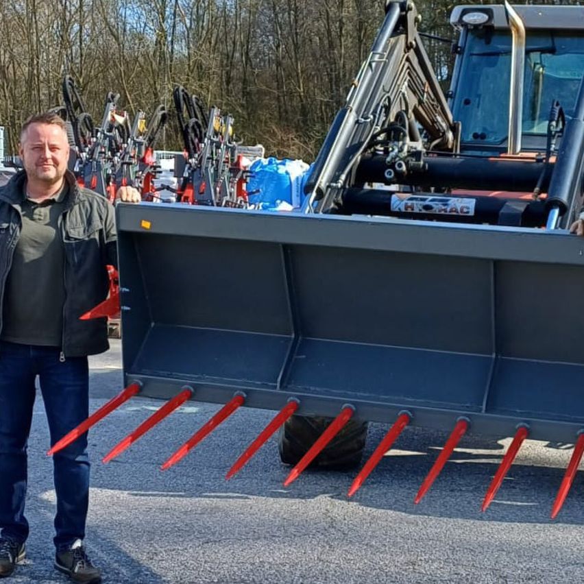 Ein Mann in einer schwarzen Jacke und Jeans steht neben einem großen, schwarzen Traktor mit roten Stacheln.