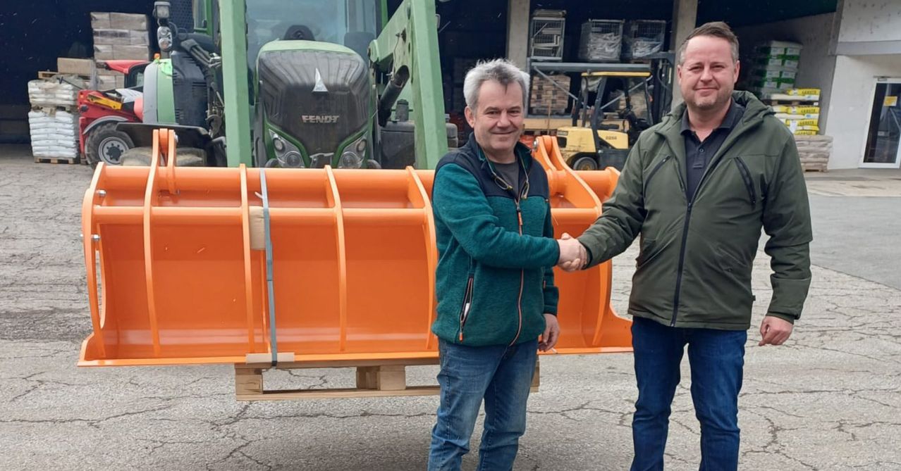 Zwei Männer schütteln sich die Hände vor einem orangefarbenen Stück Maschinerie. Der Mann auf der linken Seite lächelt. Dahinter befinden sich ein Traktor und ein Gabelstapler.