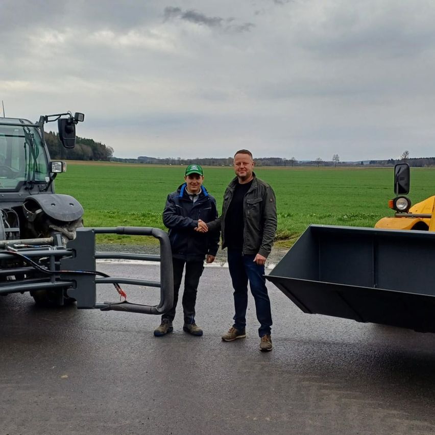 Zwei Männer stehen auf der Straße mit einem Traktor auf jeder Seite von ihnen. Sie scheinen sich die Hände zu schütteln, und der Mann auf der linken Seite trägt einen grünen Hut.