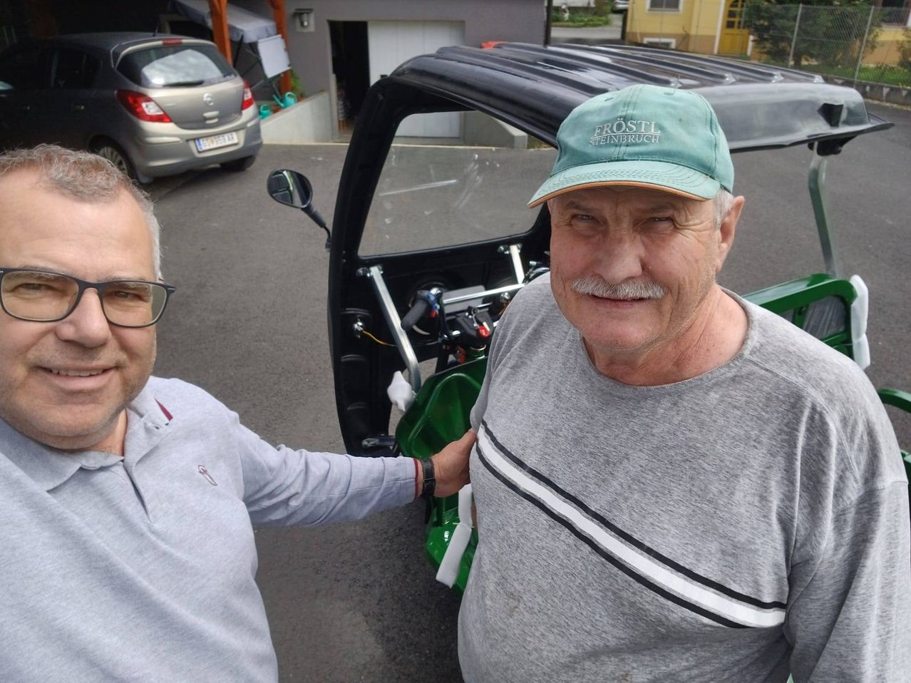Zwei Männer stehen vor einem Fahrzeug, einer trägt eine grüne Kappe mit weißem Text. Der andere trägt ein graues Shirt und eine Brille. Hinter ihnen steht ein Auto und eine Garage.