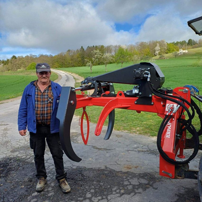 Ein Mann in einer blauen Jacke und schwarzen Hosen steht neben einem roten Traktor mit einem schwarzen Anbaugerät auf einer Landstraße.