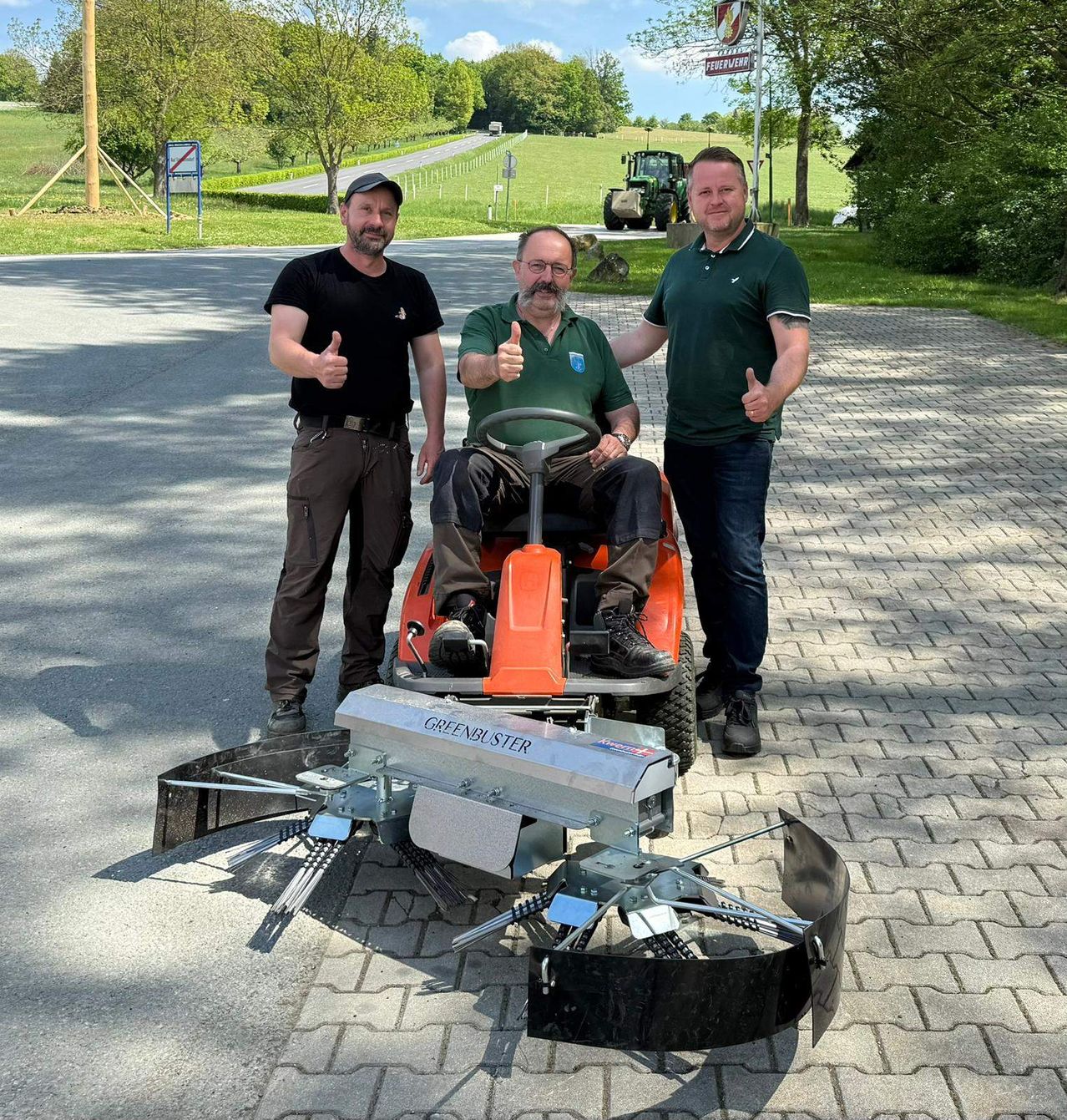 Drei Männer posieren neben einem Greenbuster-Mäher auf einer gepflasterten Auffahrt, mit einem Traktor im Hintergrund und einem Schildbrett links.