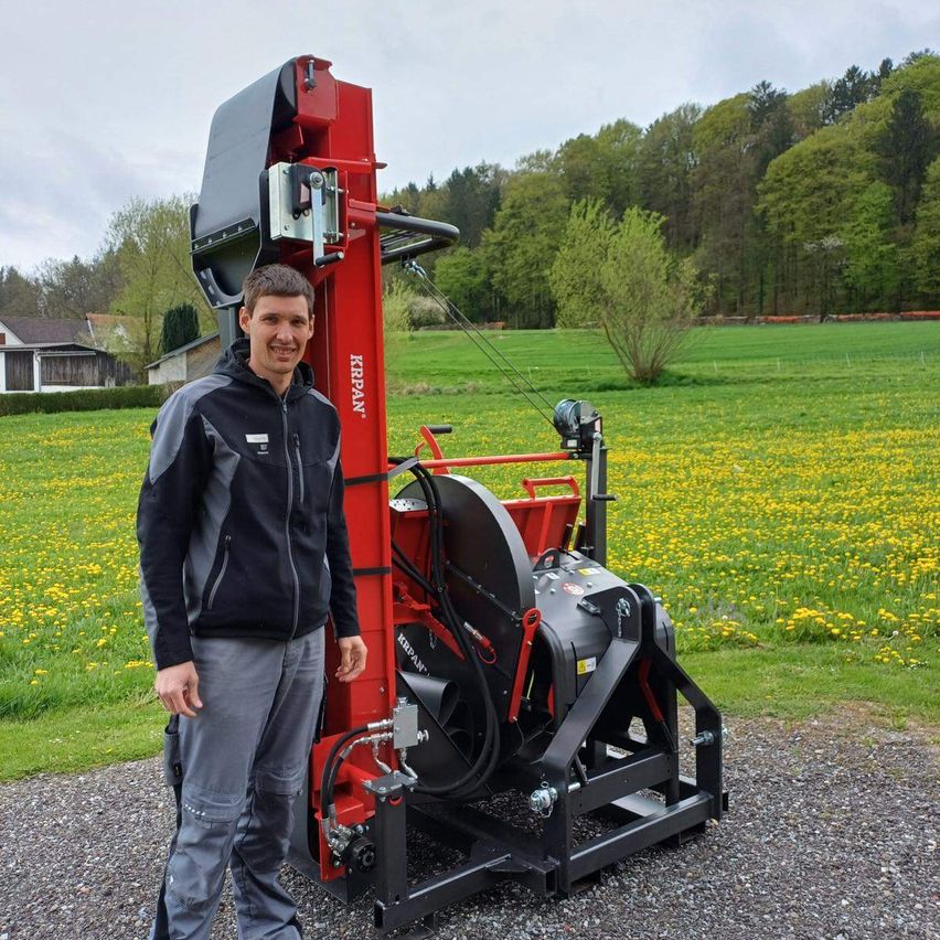 Ein Mann steht neben einer roten Maschine auf einem Kiesweg mit einem Feld gelber Blumen im Hintergrund.