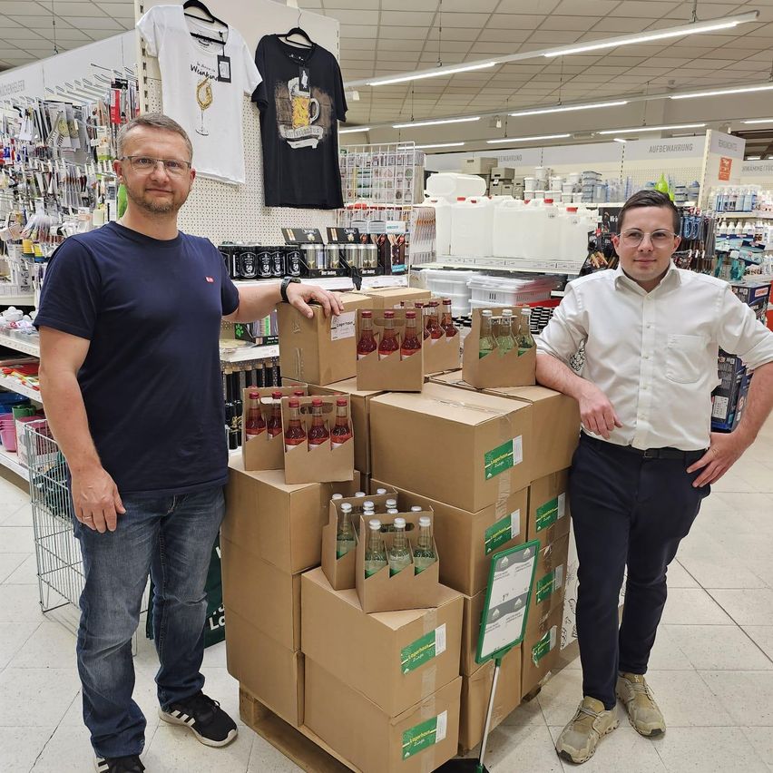 Zwei Männer stehen neben einem Stapel von Kartons mit Flaschen Getränken in einem hell beleuchteten Supermarktregal.