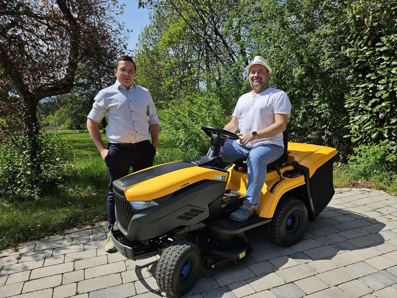 Zwei Männer stehen draußen, einer trägt einen Hut und sitzt auf einem gelben Rasenmäher, der andere in einem hellblauen Hemd und Jeans, beide lächeln.