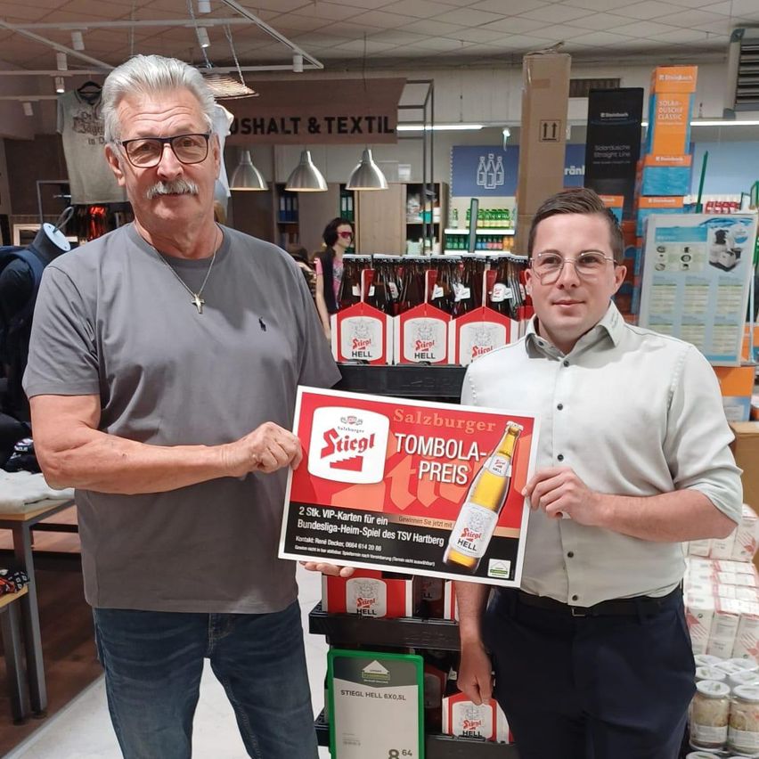 Zwei Männer stehen in einem Geschäft und halten ein Schild mit einem Flaschenbild und der Aufschrift 'TOMBOLA-PREIS'. Ein Mann trägt eine Brille, der andere eine Halskette. Hinter ihnen befinden sich Regale mit Flaschen und Schildern.